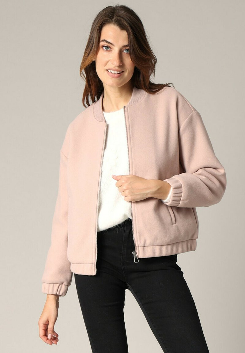 Roze fleece bomberjack met geribbelde manchetten en zoom, ritssluiting en zijzakken; gedragen over een witte trui en zwarte broek.