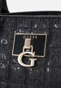 Černá texturovaná kabelka s logem, zlatými akcenty, výrazným uzávěrem a názvem značky "GUESS" na obdélníkovém panelu.