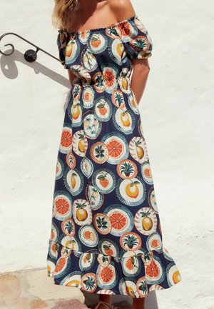 Femme portant une robe marine à épaules dénudées avec des imprimés de fruits colorés d'agrumes et d'ananas, se tenant devant un mur blanc.