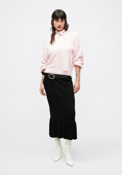 Suéter rosa claro con detalle de lazo, combinado con una falda negra acanalada con dobladillo con volantes. Botas blancas de punta puntiaguda completan el conjunto.