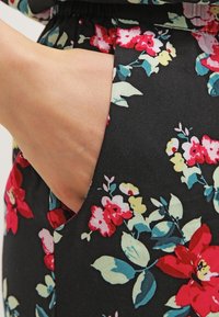 Ruka odpočívající v kapse černé květinové látky s červenými, růžovými, žlutými a tyrkysovými květy a zelenými listy.