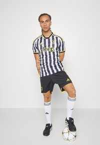 Maglia da calcio della Juventus caratterizzata da una camicia a righe bianche e nere con dettagli gialli, pantaloni neri e scarpe nere. Include un pallone da calcio.