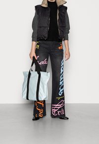 Jasnoniebieska torba tote z czarnymi uchwytami, z błyszczącym wykończeniem i zamknięciem na zamek. Noszona z ciemnymi dżinsami z kolorowymi, przerośniętymi wzorami tekstowymi.