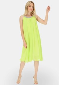 Neon żółta sukienka bez rękawów o płynnej formie, ozdobiona koralikowym dekoltem i falbaniastym wykończeniem. Zestawiona z przezroczystymi sandałami na obcasach.