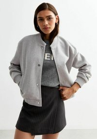 Jasnoszara kurtka bombka z napami, prążkowanym kołnierzem i rękawami, noszona na szarym swetrze z tekstem, w połączeniu z czarną spódnicą w paski.