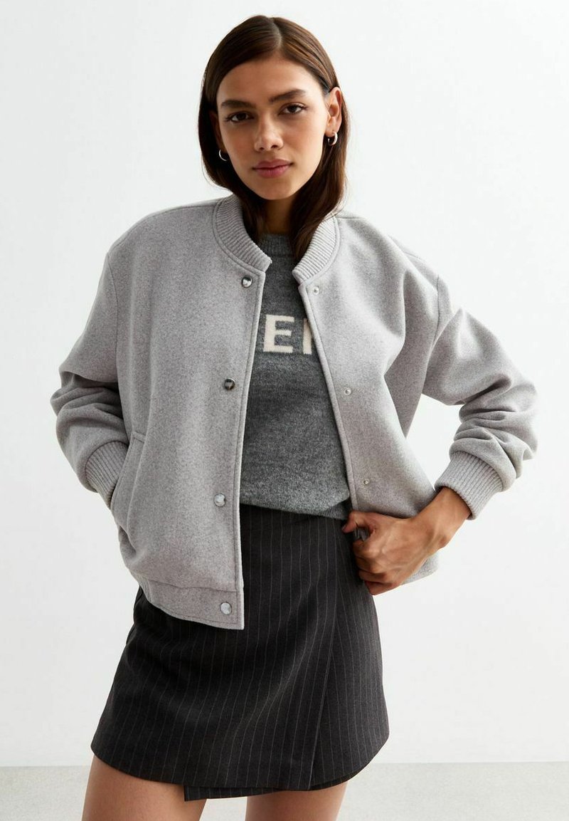 Jasnoszara kurtka bombka z napami, prążkowanym kołnierzem i rękawami, noszona na szarym swetrze z tekstem, w połączeniu z czarną spódnicą w paski.