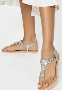 Sandali con tanga argentati decorati con strass, suola piatta color ocra e finitura lucida. Presentano un design minimalista con cinturini per un uso a dita scoperte.