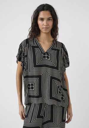 Nő fekete-fehér geometriai mintás felsőben és hozzáillő nadrágban, egyszerű szürke háttér előtt.