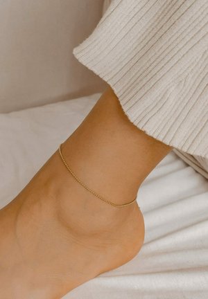 Braccialetto alla caviglia in oro con un design delicato e sottile, indossato su una caviglia nuda; pelle liscia con uno sfondo in tessuto bianco texturizzato.