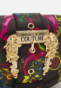Bolso colorido con una superficie negra texturizada y patrones florales en rosa y amarillo. Herrajes dorados con la etiqueta "Versace Jeans Couture".