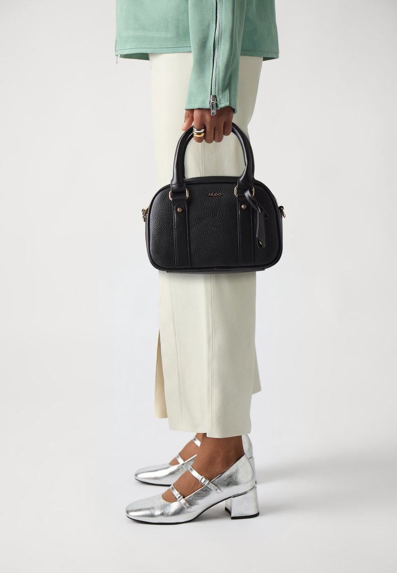 Personne tenant un sac à main en cuir noir, portant une veste verte, une jupe crème, des chaussures à talons carrés argentés et des bagues en or aux doigts.
