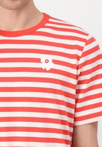 Tricou cu dungi roșii și albe, cu guler rotund, având un mic logo alb în formă de floare pe partea stângă a pieptului. Fabricat din material moale.
