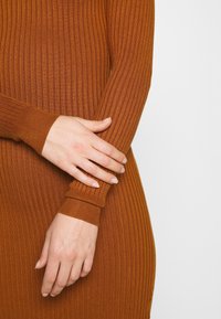 Mains reposant sur une robe tricotée couleur rouille à manches longues côtelées, sur un fond clair uni.