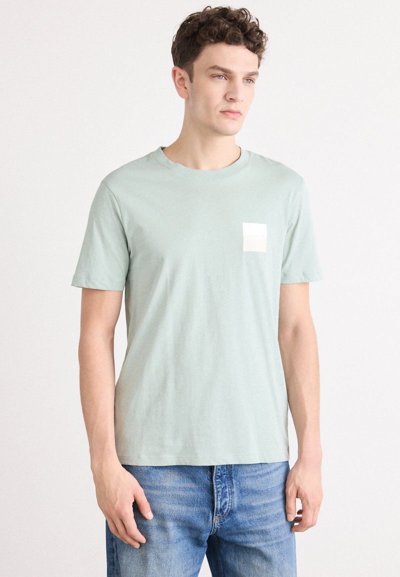 Camiseta de algodón verde claro con cuello redondo, mangas cortas y un pequeño parche de logo blanco en el lado izquierdo del pecho. Se lleva con vaqueros de mezclilla azul.
