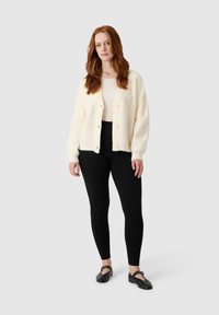 Kremowy sweter z dzianiny z prążkowanymi detalami i guzikami, zestawiony z czarnymi legginsami i płaskimi czarnymi butami. Prosty, teksturowany design.