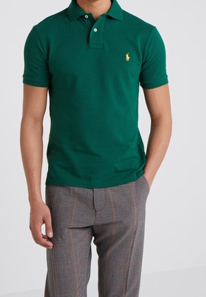 Mand iført en grøn kortærmet poloshirt med et gult logo og grå ternede bukser, står med den ene hånd i lommen.