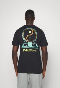 Hombre con una camiseta negra que tiene un gráfico de una canasta de baloncesto y una pelota en colores verde neón y amarillo en la parte de atrás, combinado con pantalones de chándal grises.