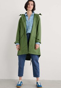 Veste imperméable verte, légère, avec une capuche, des boutons-pression et des poches, associée à une chemise en denim bleu et un jean court bleu.