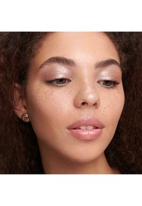Gros plan sur un visage avec une peau bronzée parsemée de taches de rousseur, des lèvres nues brillantes et un maquillage des yeux scintillant. Des boucles d'oreilles créoles dorées sont portées.