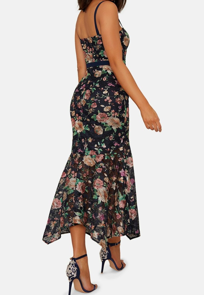 Femme portant une robe noire moulante à motifs floraux avec des parties transparentes et des talons hauts bleu marine décoratifs, tournée de dos avec le bras droit légèrement plié.