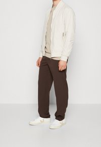 Jasnobeżowa kurtka bomber, beżowy top, ciemnobrązowe spodnie i białe trampki z beżowymi akcentami. Gładka tekstura, prosty design.