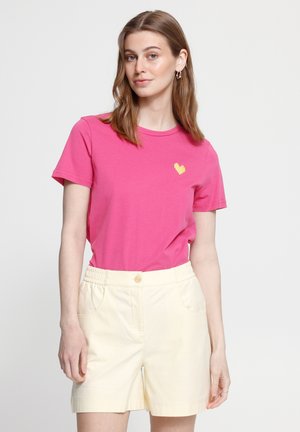 Femme vêtue d'un t-shirt rose avec un petit cœur jaune sur la poitrine et un short taille haute couleur crème, debout devant un fond uni.