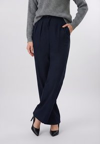 Γυναίκα που φοράει σκούρο ναυτικό παντελόνι με φαρδιά πόδια και πτυχώσεις, γκρι ριμπ πουλόβερ, μαύρα μυτερά ψηλοτάκουνα, στέκεται με το ένα χέρι στην τσέπη.