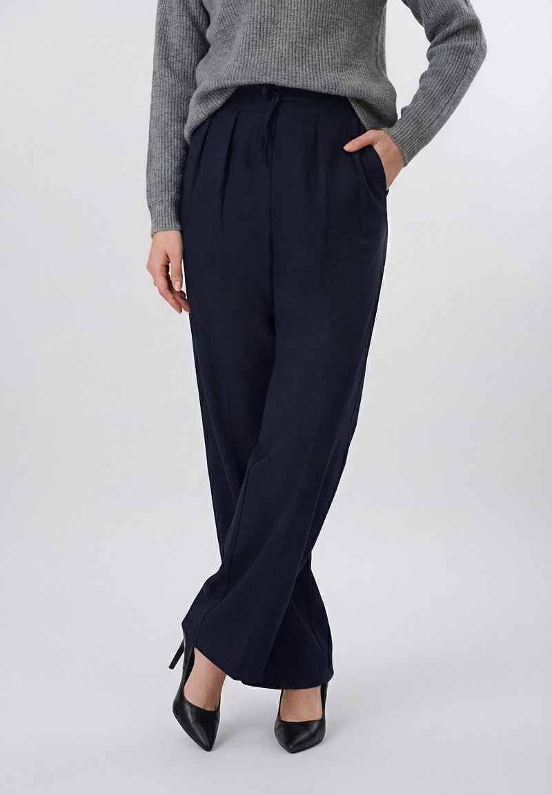 Γυναίκα που φοράει σκούρο ναυτικό παντελόνι με φαρδιά πόδια και πτυχώσεις, γκρι ριμπ πουλόβερ, μαύρα μυτερά ψηλοτάκουνα, στέκεται με το ένα χέρι στην τσέπη.