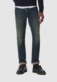 Jeans de mezclilla azul oscuro con corte regular, que presentan un ligero efecto de desvanecimiento y dobladillos enrollados. Llevados con zapatos de cuero negros.