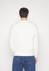 Sudadera blanca con cuello redondo, mangas largas y puños acanalados. Textura suave, diseño minimalista, combinada con jeans de mezclilla azul.