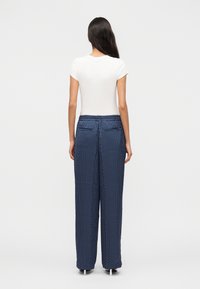 Γυναίκα με μακριά σκούρα μαλλιά στέκεται γυρισμένη με την πλάτη, φορώντας λευκό κοντομάνικο τοπ και μπλε σκούρο παντελόνι με φαρδιά πόδια και λευκές πουά βούλες.