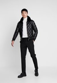 Zwarte gewatteerde leren bomberjack met een nepbontkraag, gedragen over een witte coltrui, in combinatie met zwarte broeken en schoenen.