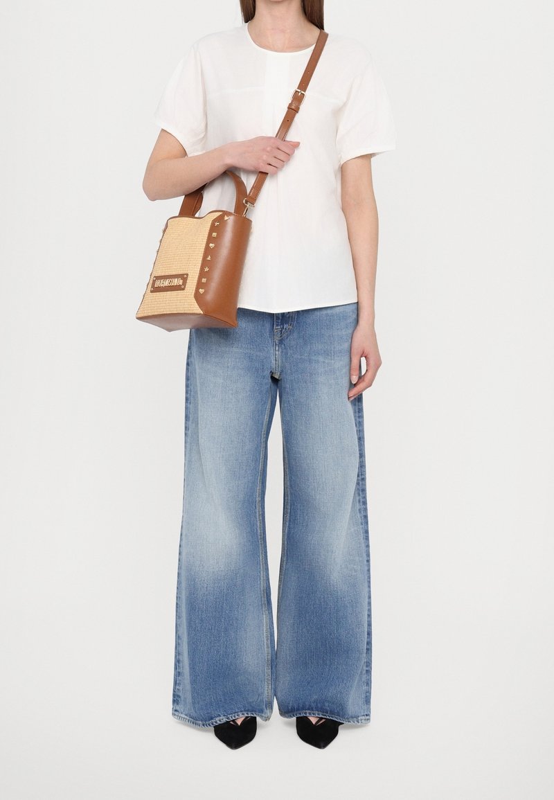Blouse blanche à manches courtes, jean bleu clair à jambes larges, et un sac à main marron tissé avec des clous et une bandoulière. Chaussures noires à bout pointu.