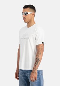 Tricou alb din bumbac cu guler rotund, având un logo gri deschis pe față și un eticheta neagră pe mânecă. Modelul poartă ochelari de soare.