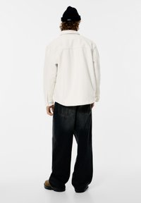 Valkoinen fleecetakki, väljä mustat denimhousut, musta neulottu pipo, näkyvillä ompeleilla ja taskuilla, esitetty takaa.