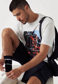 Camiseta blanca con un diseño gráfico en azul, rojo y negro, combinada con pantalones cortos negros y calcetines con patrón. Zapatillas blancas y una mochila negra.