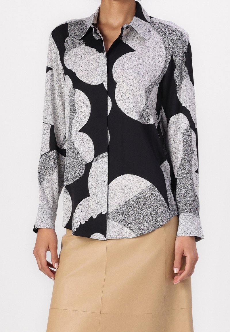 Femme portant une chemise boutonnée à manches longues avec un motif abstrait noir et blanc et une jupe beige.