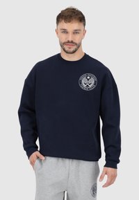 Felpa navy con scollatura a girocollo, maniche lunghe e logo frontale. Il materiale sembra morbido; la vestibilità è rilassata con uno stile casual.