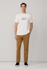 Mann steht mit einem weißen Grafik-T-Shirt, braunen Hosen und schwarzen Turnschuhen vor einem schlichten, hellgrauen Hintergrund.