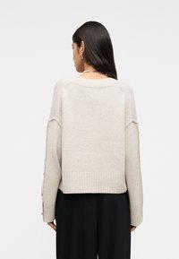 Femme aux longs cheveux noirs portant un pull en maille beige avec des boutons sur les manches et un pantalon noir à jambes larges, vue de dos.