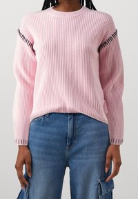 Rosa stickad tröja med ribbad struktur, rund halsringning och kontrasterande svart sömnad längs ärmöppningarna. Matchad med blå jeans.