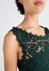 Grön spetsklänning med blommönster, genomskinlig överdel vid halsringningen och detaljerad brodering. Slät textur, ärmlös design.