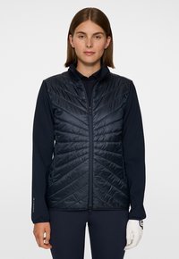 Kvinna iklädd en svart quiltad väst över en svart långärmad tröja, med en vit golfhandske på sin högra hand, vänd framåt.