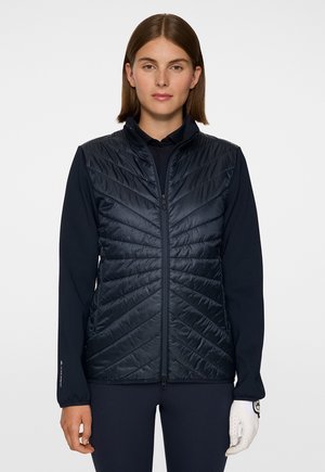 Frau trägt eine schwarze gesteppte Weste über einem schwarzen Langarmtop, mit einem weißen Golfhandschuh an ihrer rechten Hand, schaut nach vorne.