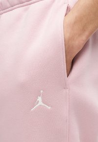 Χέρι στην τσέπη ροζ ανοιχτόχρωμου υφασμάτινου παντελονιού με μικρό λευκό λογότυπο Jordan Jumpman κοντά στη ραφή της τσέπης.