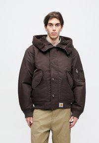 Καφέ μόνωτη ζακέτα με υφασμάτινη, σκούρα καφέ κουκούλα. Διαθέτει μεγάλες μπροστινές τσέπες, μια τσέπη με φερμουάρ στο μανίκι και μια ετικέτα με λογότυπο.