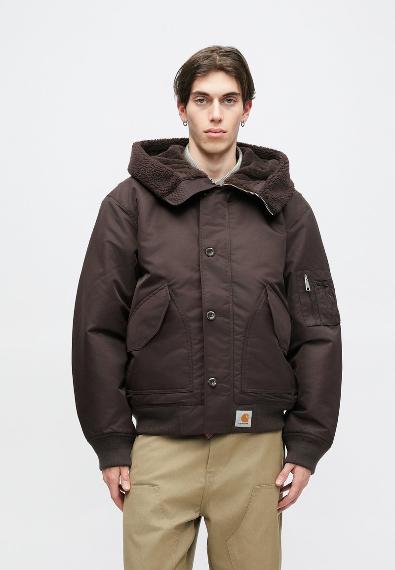 Καφέ μόνωτη ζακέτα με υφασμάτινη, σκούρα καφέ κουκούλα. Διαθέτει μεγάλες μπροστινές τσέπες, μια τσέπη με φερμουάρ στο μανίκι και μια ετικέτα με λογότυπο.