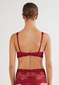 Czerwony koronkowy biustonosz z regulowanymi ramiączkami, z falbaniastym brzegiem i częściowo przezroczystymi elementami, prezentujący misterny wzór kwiatowy.