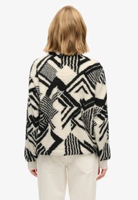 Fuzzy pullover jacket in cream with black geometric patterns, featuring a round neckline and ribbed cuffs, paired with light-colored pants.