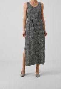 Femme portant une robe maxi sans manches noire avec un imprimé floral blanc et une fente sur le côté, accompagnée de chaussures à talons gris.